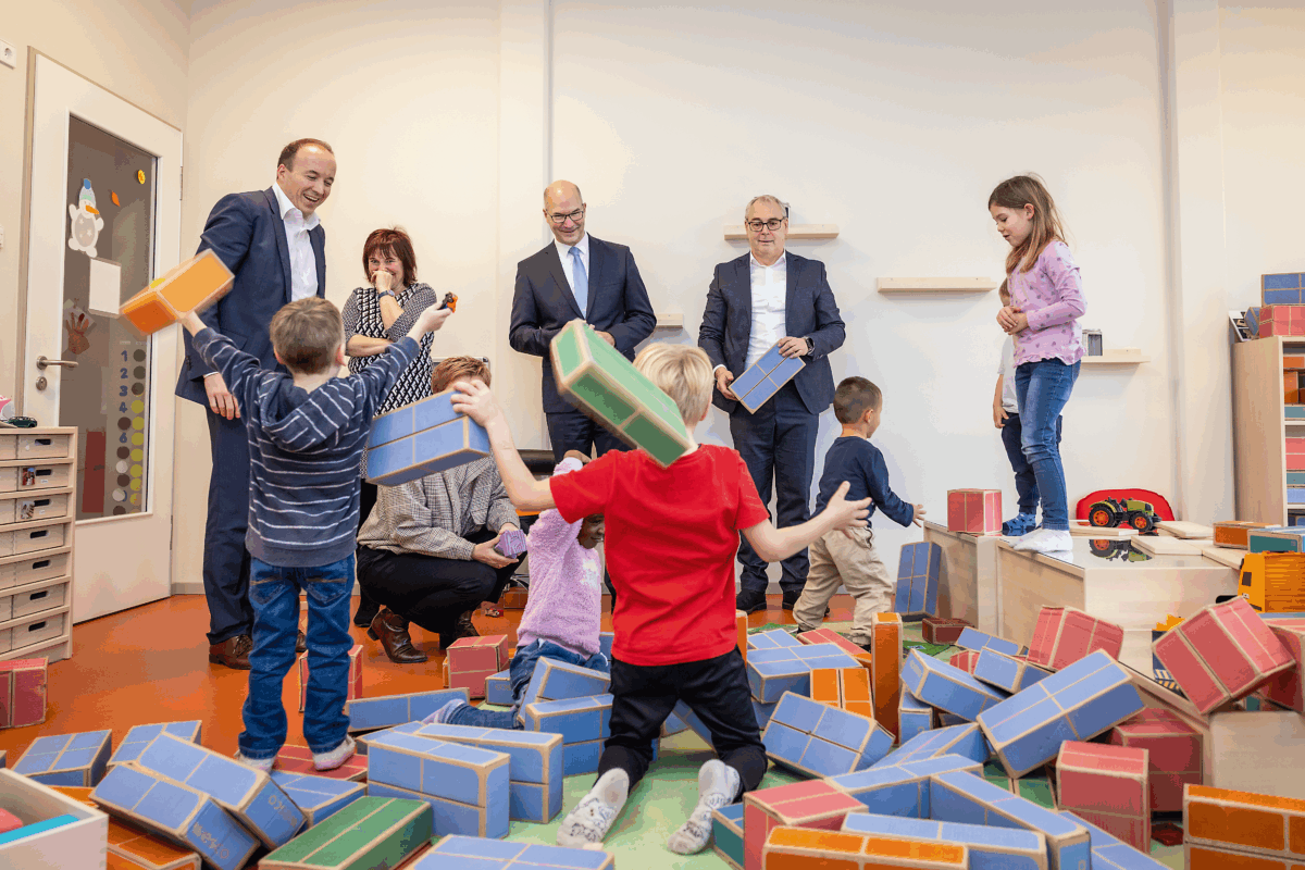 Kinder spielen mit Holzklötzen. Besucher schauen zu.