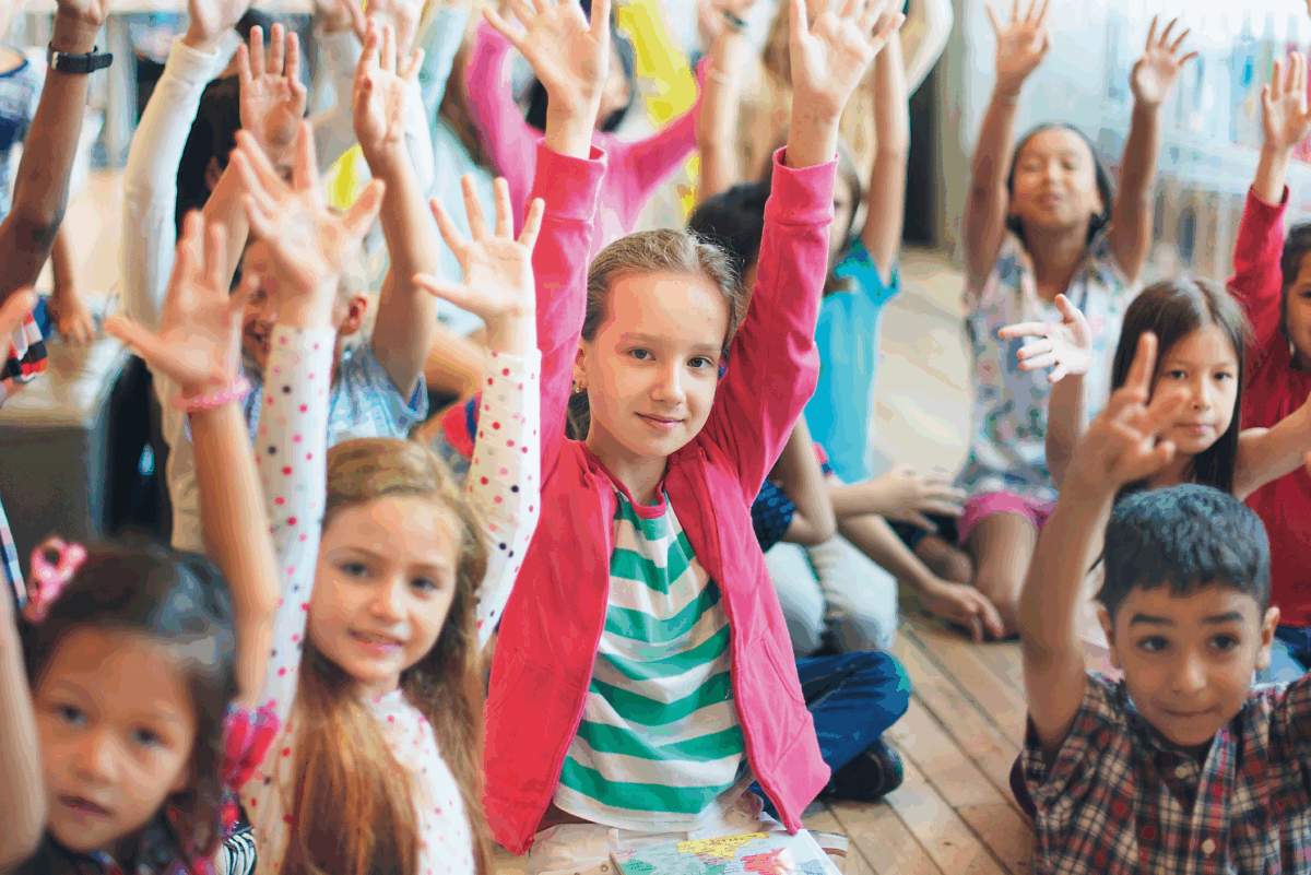 Schulkinder sitzen auf dem Boden und strecken beide Hände in die Luft.
